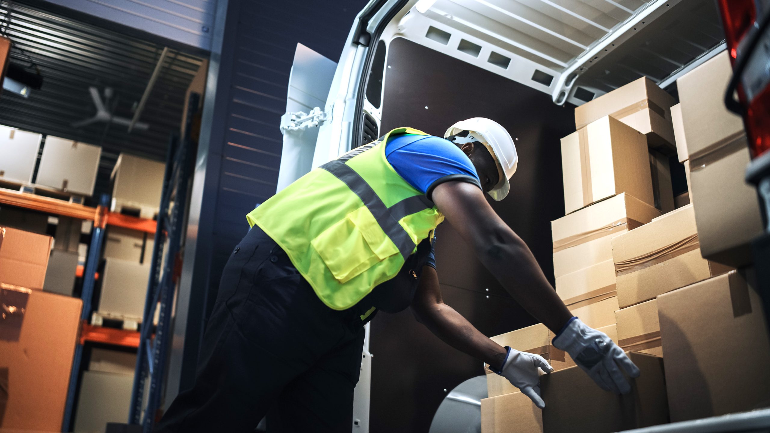 Handsome Black Male Retail Warehouse Worker  Loads Cardboard Boxes in  Delivery Truck. Logistics Retail Center, Delivering e-Commerce Online Orders, Food and Medicine Supply. Low Angle Evening Shot